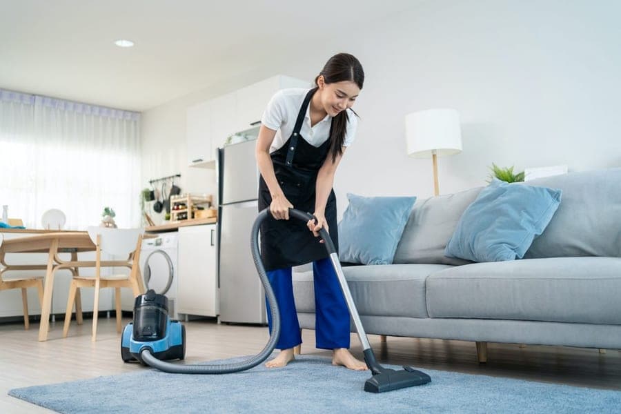 Femme en tablier noir qui passe l'aspirateur sur un tapis bleu clair dans un séjour ouvert sur la cuisine