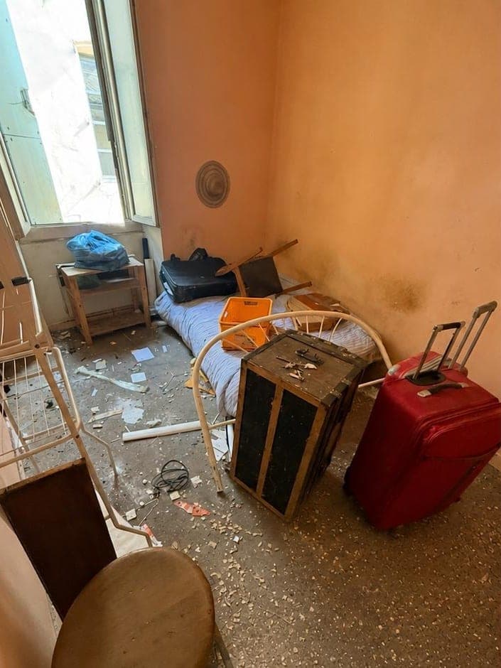 Petite chambre aux murs saumon avant intervention : sommier, valise, malle, chaise renversée et gravats au sol